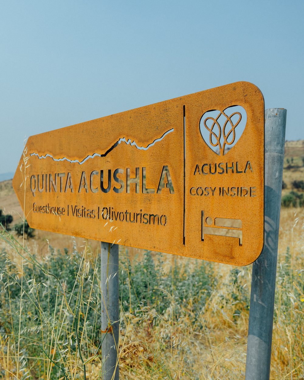 Wegweiser aus Cortenstahl mit der Aufschrift „Quinta Acushla — Guesthouse, Visitas, Olivoturismo“ am Feldweg zur Farm